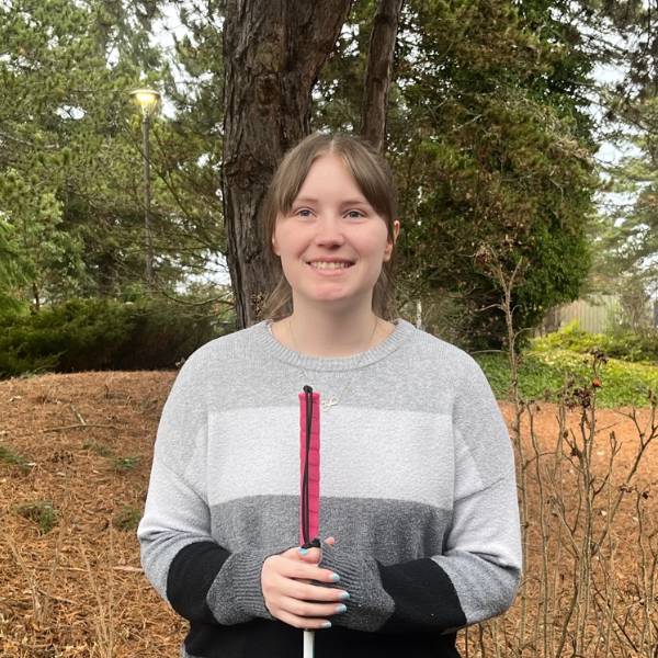 A headshot of Jordan. She smiles against a wooded background, wearing a gray and black striped sweater and holding a walking stick..
